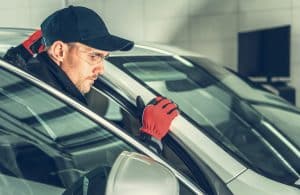 A man wearing safety glasses, a cap, and red gloves inspects the frame of a silver car inside a well-lit garage, focusing on auto glass repair to ensure the vehicle’s safety and integrity.