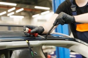 A person wearing black gloves uses a screwdriver to work on the roof area of a car, possibly removing or installing a weatherstrip or trim as part of an auto glass replacement, with wires visible inside the car.