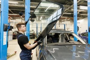 A mechanic wearing gloves lifts a large windshield in an auto repair shop, preparing for glass replacement on a gray vehicle. The bright workshop features blue support beams and other cars in the background.