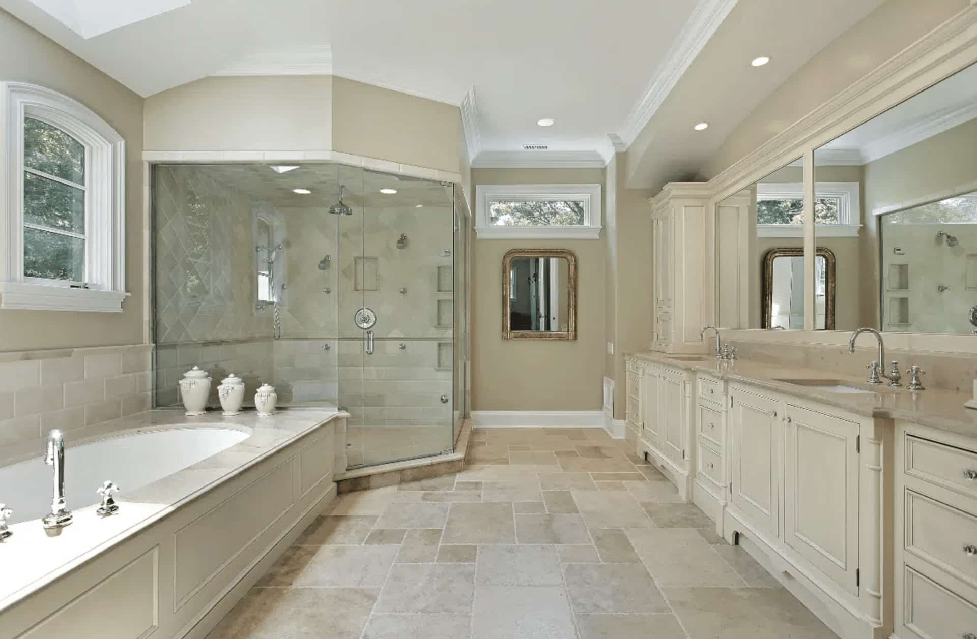 Spacious, elegant bathroom with large glass shower, built-in bathtub, cream cabinetry, double sinks, beige tile floor, and two windows bringing in natural light.